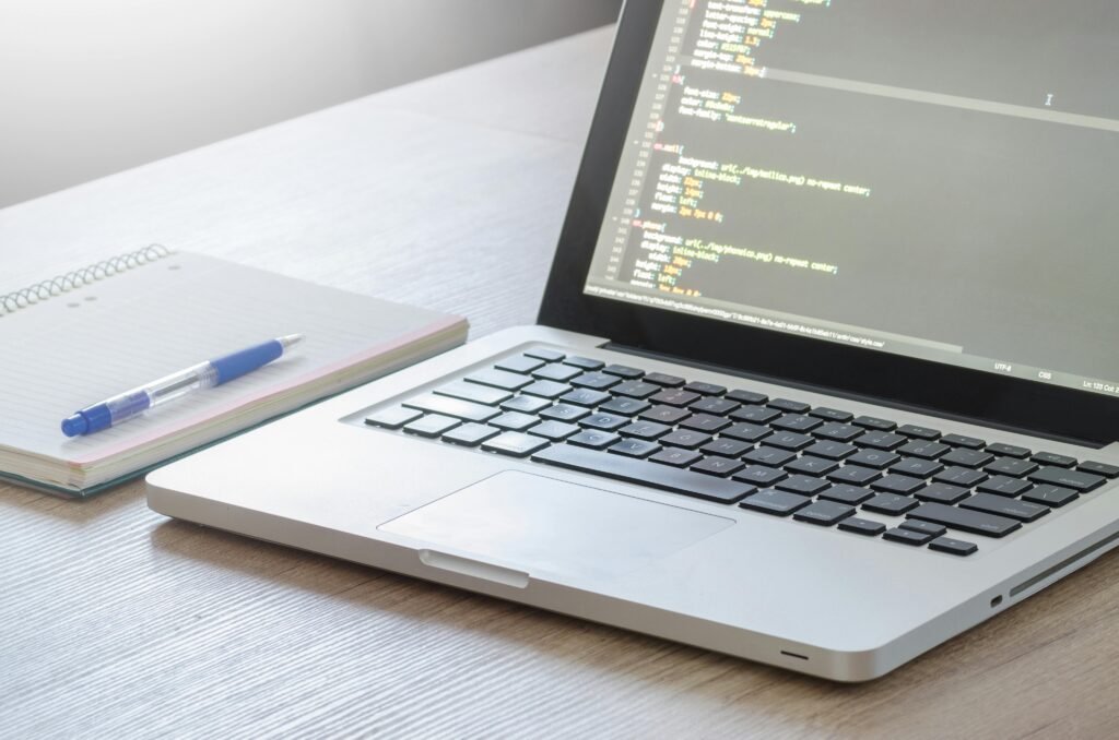 pexels-photo-574073-574073 Laptop with code editor open, notebook and pen on wooden desk, perfect for tech and work themes.